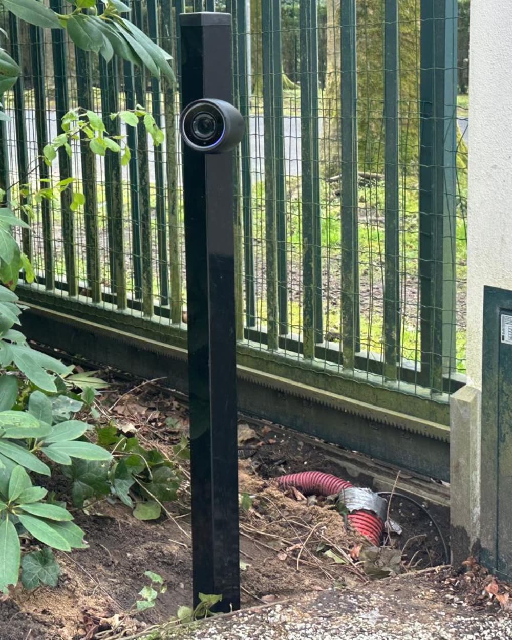 Beveiligingscamera op een zwarte paal langs een hekwerk.
