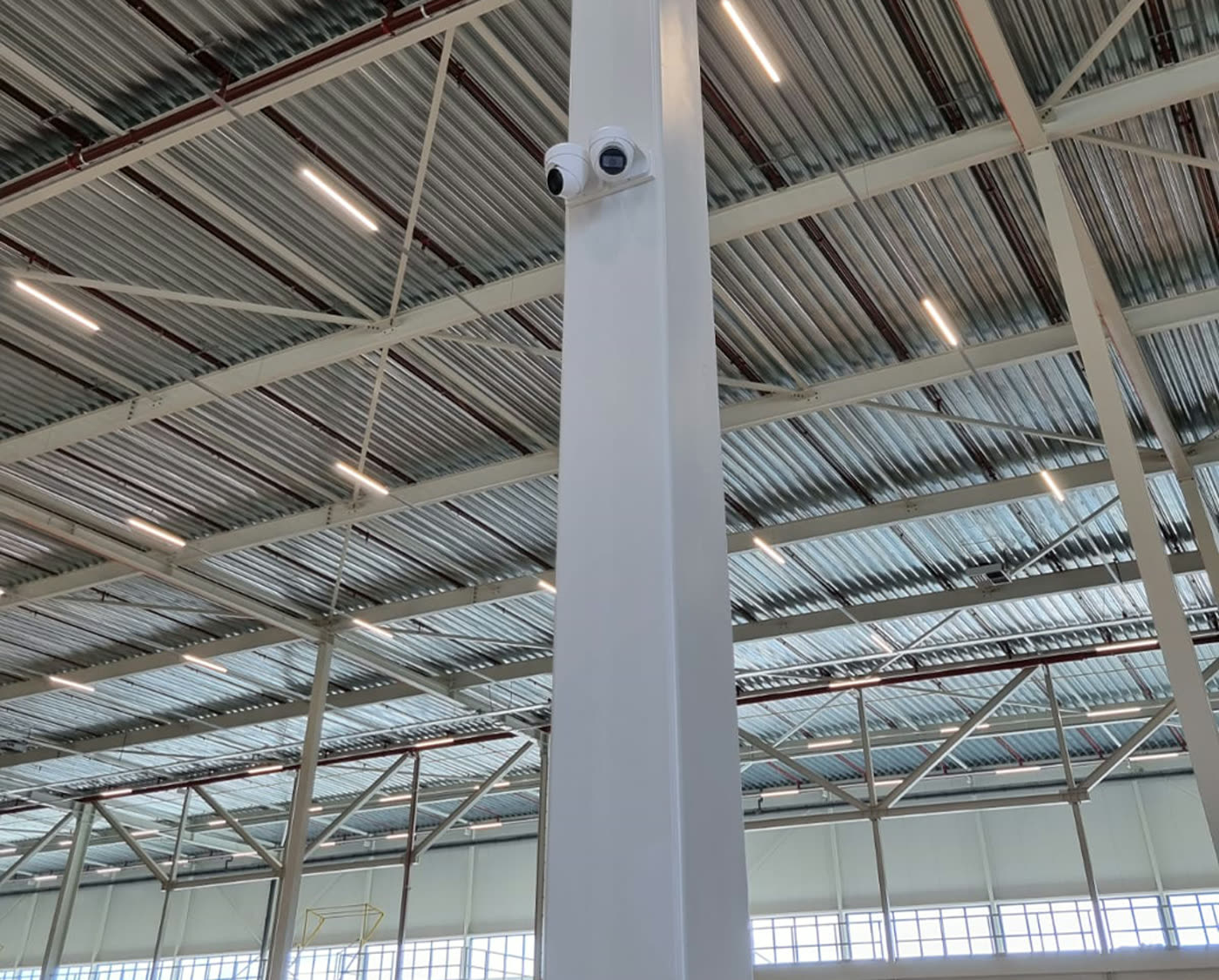 Twee beveiligingscamera’s op een stalen kolom in een grote bedrijfshal met ledverlichting in het dak.