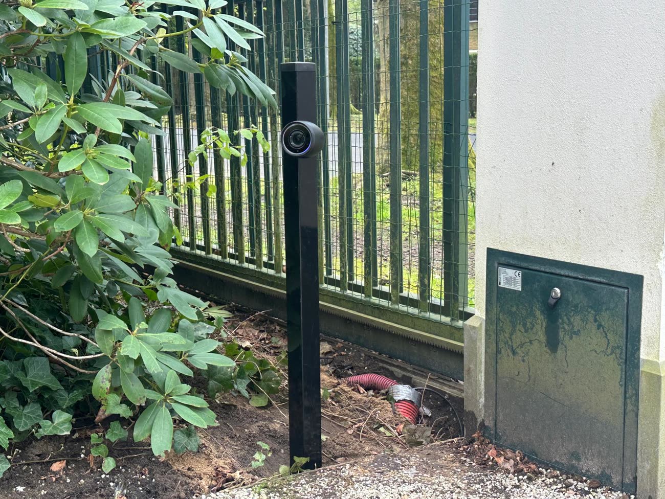 Camera op een zwarte paal bij een groen hekwerk, gericht op de oprit.