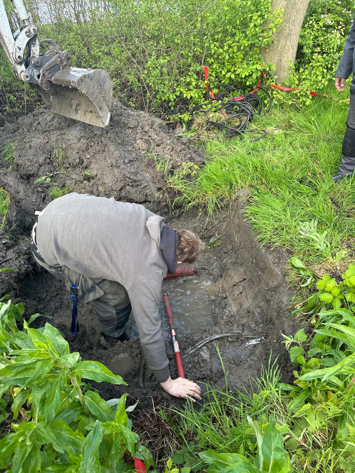 Monteur graaft een beschadigde kabel vrij in een natte sleuf naast een minigraver.
