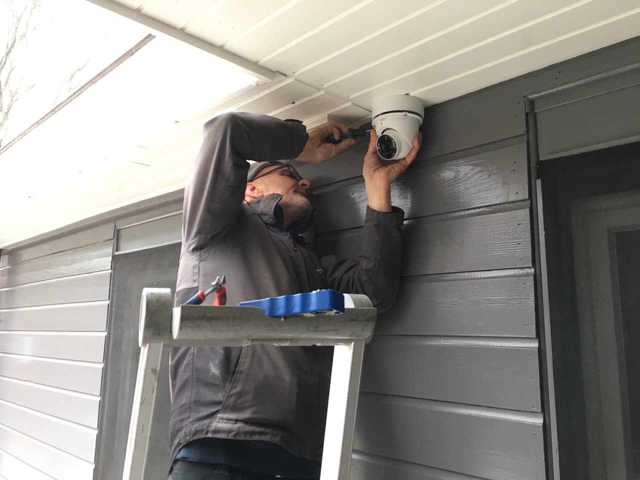 Monteur monteert een witte camera onder de dakrand aan de gevel van een woning.