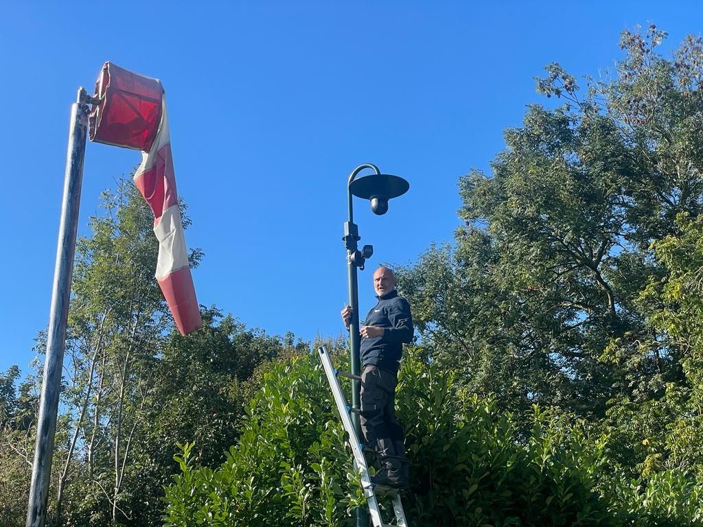 Monteur op een ladder bij een beveiligingscamera en een windzak op een buitenterrein.