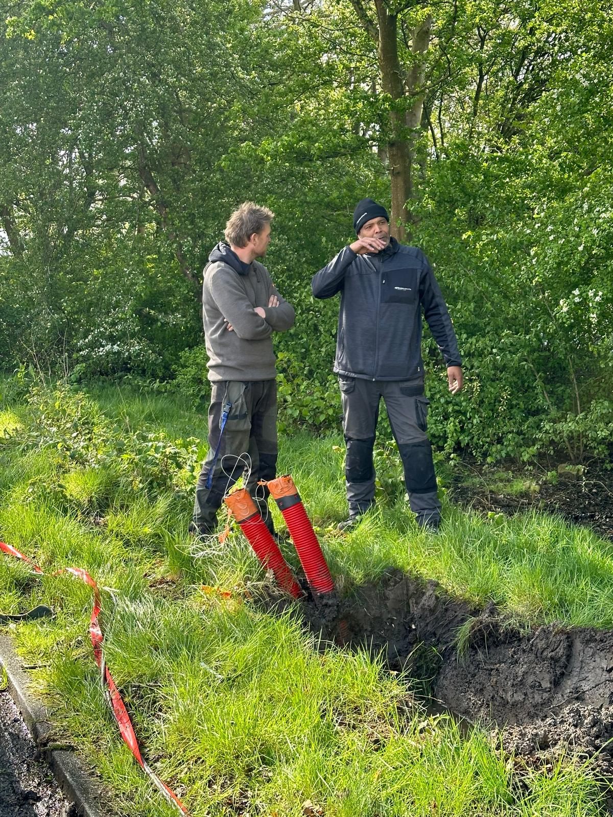 Twee monteurs overleggen bij een open sleuf met rode mantelbuizen voor kabels.