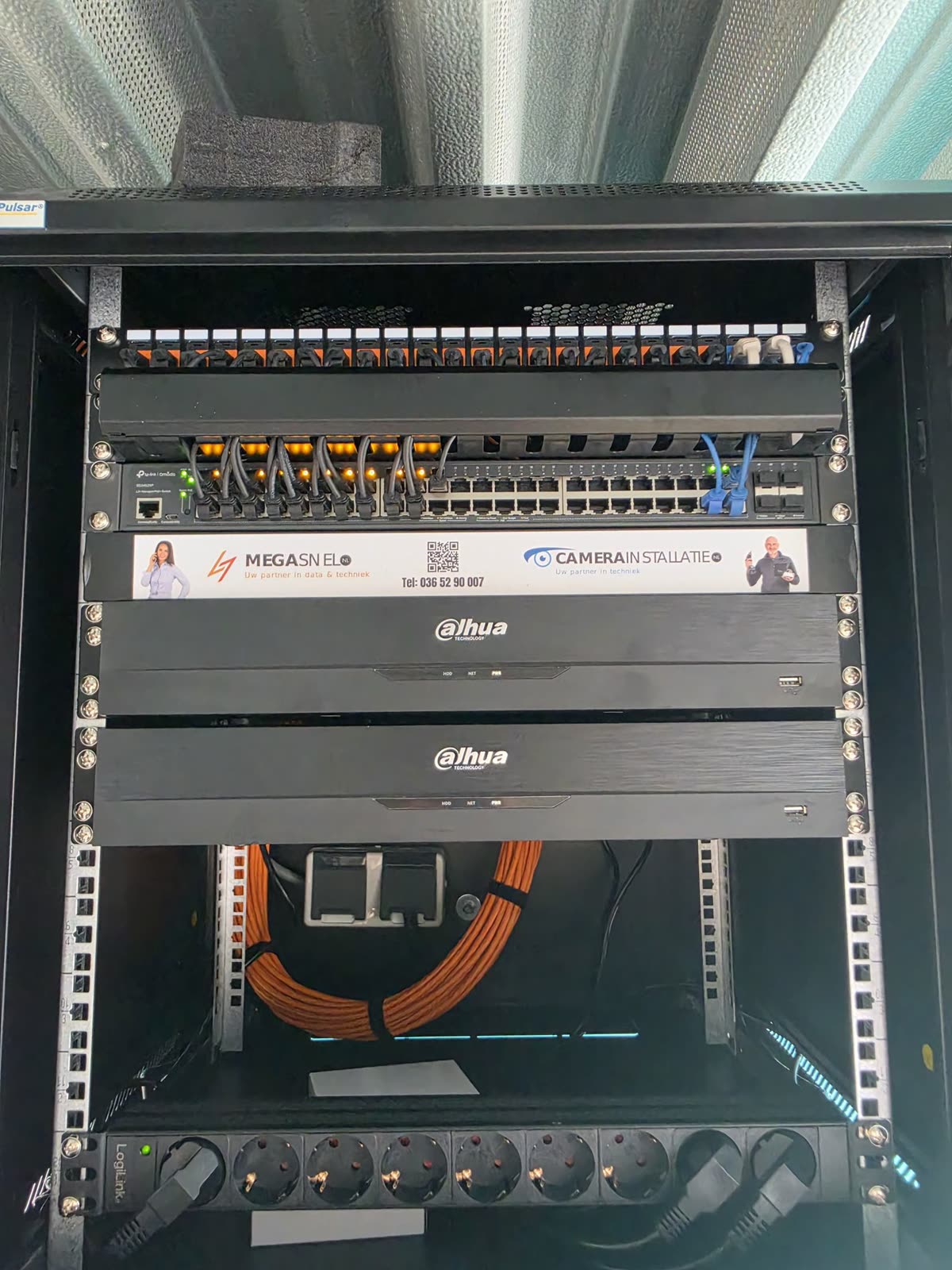 Geopende patchkast met patchpaneel, PoE-switch en twee netwerkvideorecorders, aangelegd met oranje datakabel.