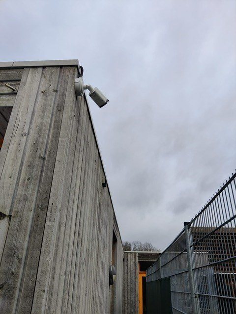 Camera op de hoek van een houten gevel, gericht langs het hekwerk.