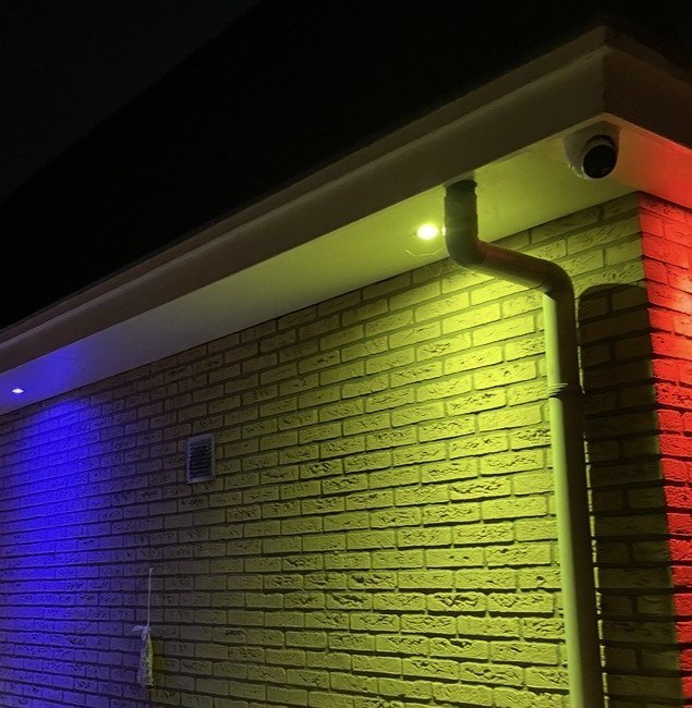 Gevel in de avond met gekleurd licht van inbouwspots in de overstek, en een camera onder de dakrand.