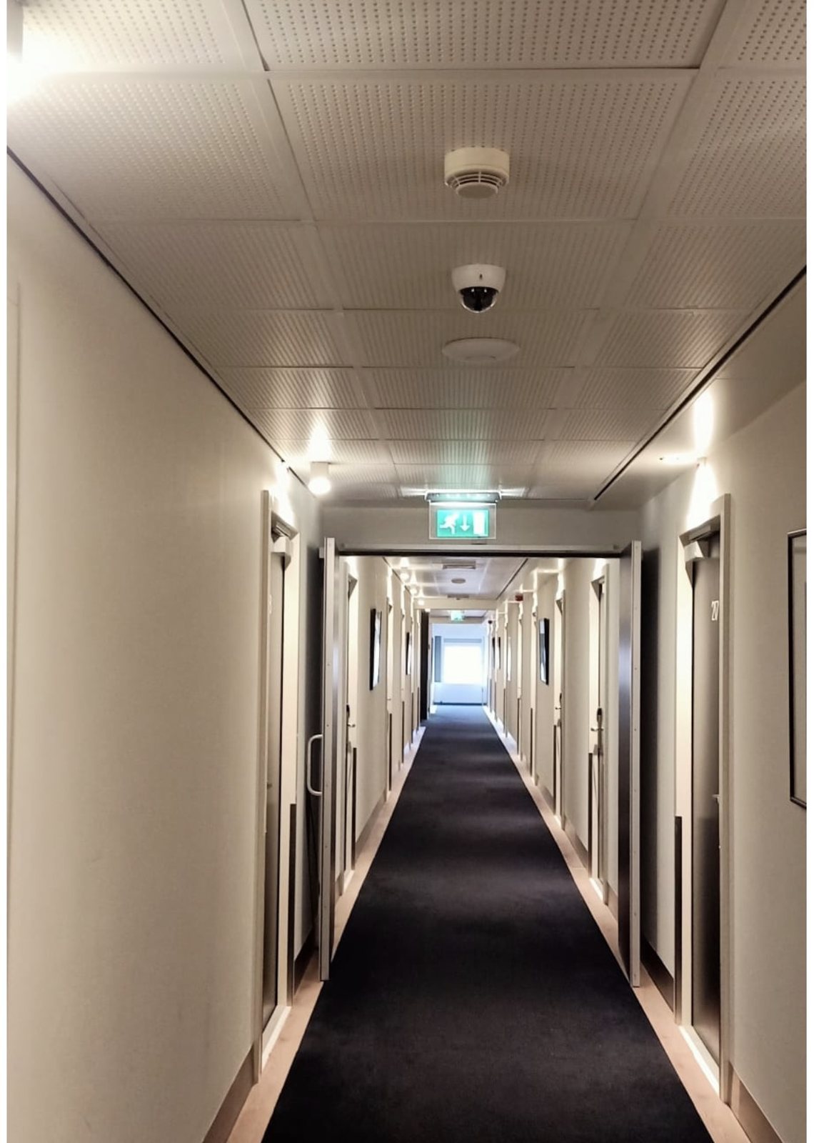 Hotelgang met een verlicht nooduitgangbord, een rookmelder en een domecamera in het plafond.