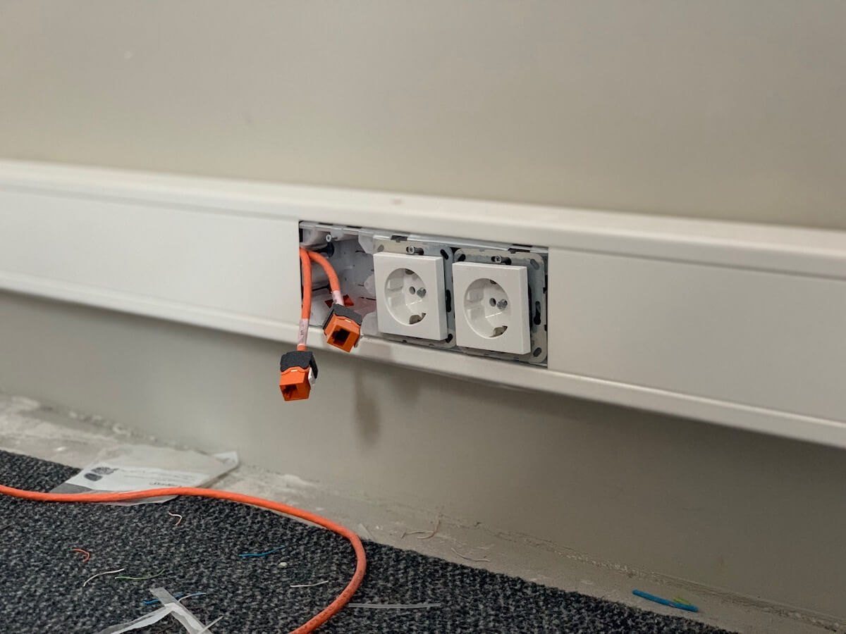 Kabelgoot langs de wand met stopcontacten en oranje netwerkaansluitingen.