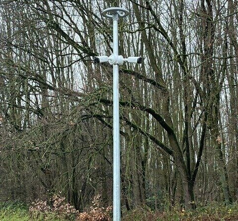 Mast aan de terreinrand met verlichting bovenin en twee camera's.