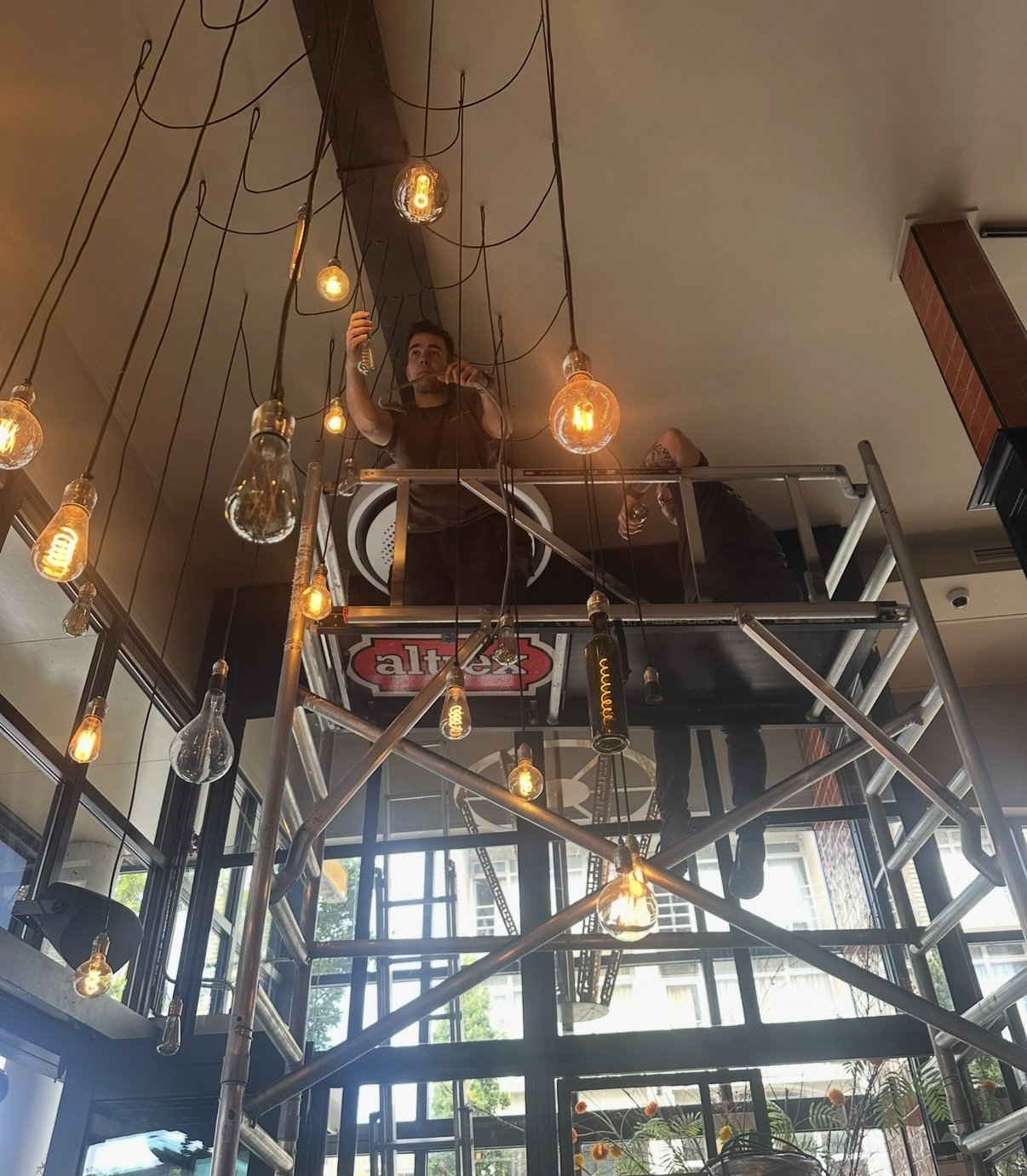 Twee monteurs op een rolsteiger hangen tientallen hanglampen met filamentlampen op in een horecazaak.