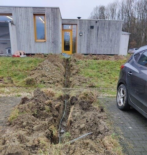 Open sleuf met kabel richting een houten bijgebouw op het terrein.