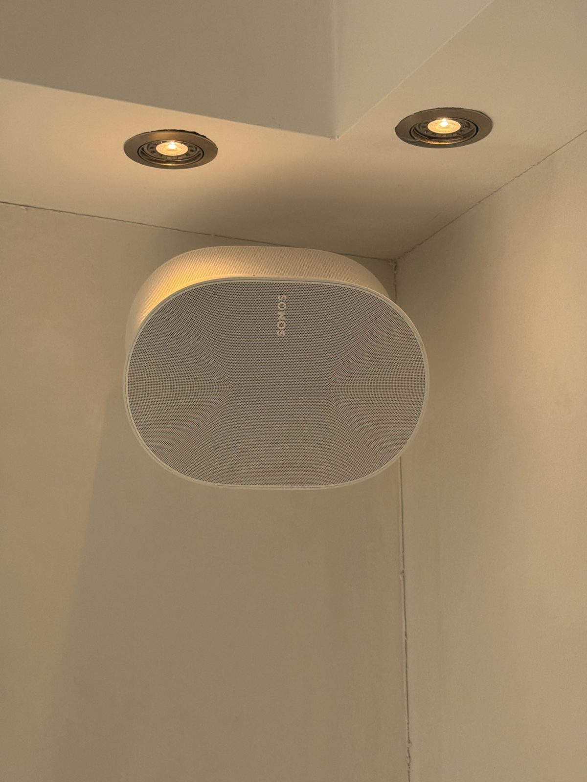 Witte wandspeaker in de hoek van een kamer, onder twee inbouwspots.